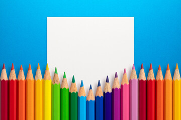 Creative layout made of color pencils on blue paper background with white paper card note. Flat lay. Education concept.