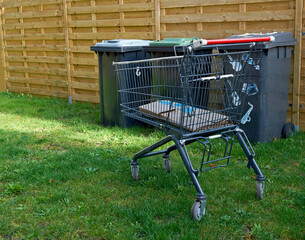 A taken-away shopping cart in a garden. In the background are large garbage containers