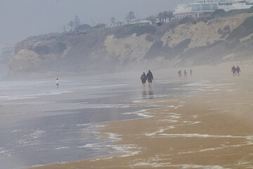 Paisaje personas caminando por la playa en otoño con niebla y mucho viento en Conil de la Frontera,  Cádiz