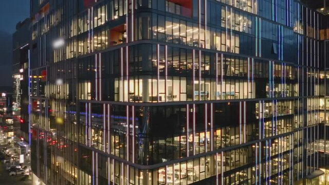 Drone shot at a night skyscraper with glowing office windows and business people working late at computers. Flyby of an office building, panorama to the left