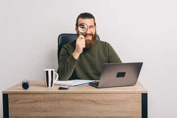 Cheerful bearded male worker is sitting in the office while looking through a magnifying glass.
