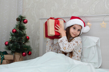 Christmas morning, a little girl in pajamas in bed rejoices with gifts on the background of a Christmas tree. A happy, smiling child opens a New Year's gift at home. The concept of holidays, Christmas