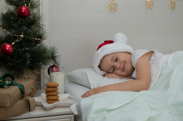 Obraz na płótnie Canvas Christmas cookies and a glass of milk are a traditional treat. A charming little boy fell asleep on the bed near the bedside table with a treat for Santa Claus. The concept of the New Year. Relatives.