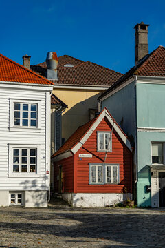 kleines, altes rotes Haus mit weissen Fensterrahmen in Bergen, Norwegen, das zwischen grossen neueren H&auml;usern in der Altstadt steht. herziges, echt nordisches H&auml;uschen mit Stil. norwegischer Flair 