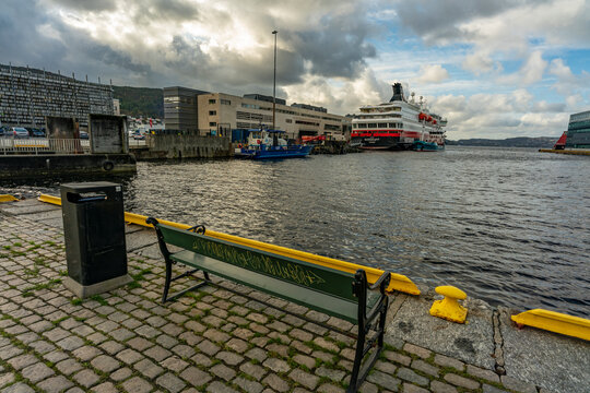 Bank im Hafen von Bergen, mit Blick auf die Anlegestelle, Dock und Kai mit Schiffen und den angrenzenden Wirtschaftsgebäuden, Speicher und Handelshaus. gelb grüne Parkbank