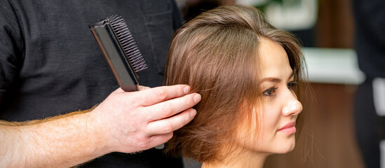 Male hairdresser works on the hairstyle of the young caucasian brunette woman at a hair salon