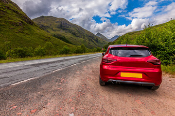 Naklejka premium Mountains in the Scottish Highlands. Road through the countryside with a red car in the foreground. Clouds in the sky in the daytime with sunshine. Green trees and bushes