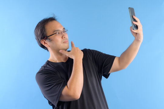 Asian man wore glasses and wear black t-shirts. Using mobile To take a picture  or selfie of himself isolated on blue background.