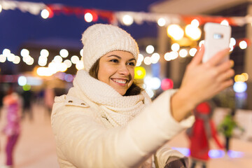 Christmas, winter, technology and leisure concept - happy young woman taking picture with smartphone on ice skating rink outdoors.