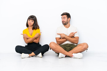 Obraz premium Young couple sitting on the floor isolated on white background pointing to the laterals having doubts