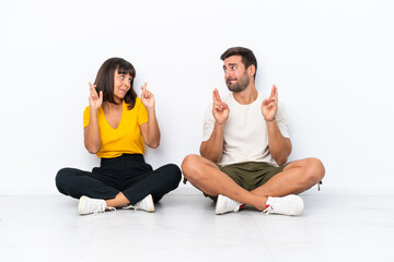 Fototapeta na wymiar Young couple sitting on the floor isolated on white background with fingers crossing and wishing the best
