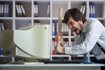 Angry man is destroying old computer with baseball bat in office.