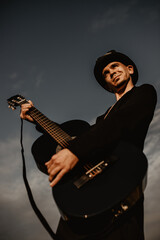 Young man plays the guitar in the field. He is wearing a black coat, hat and glasses. It's cloudy outside