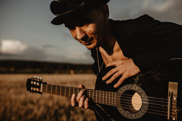 Young man plays the guitar in the field. He is wearing a black coat, hat and glasses. It's cloudy...