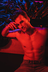 sexy shirtless man looking at camera near black bedding in red lighting