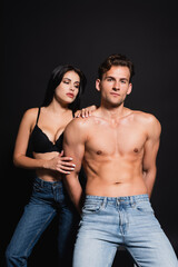 Fototapeta na wymiar sexy shirtless man in jeans looking at camera near sensual brunette woman isolated on black