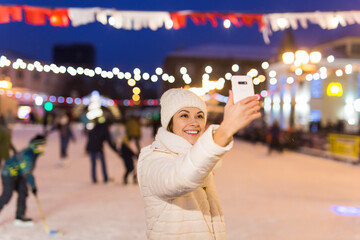 Happy young woman in winter on the ice rink taking picture on smartphone, selfie.