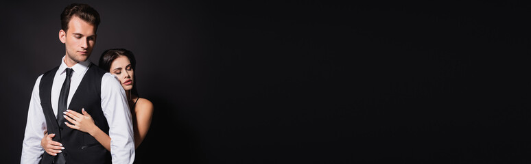 sensual woman with closed eyes hugging stylish man isolated on black, banner