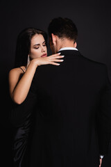 back view of man in suit near elegant woman hugging him isolated on black