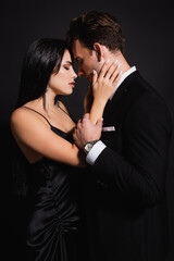 brunette woman in silk dress touching face of elegant man isolated on black