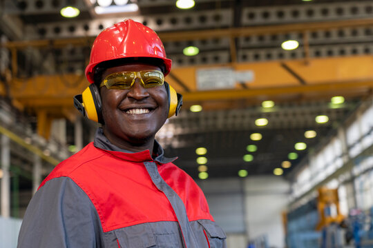 Smiling Young African American Worker In Personal Protective Equipment Looking At Camera - Portrait Of Black Industrial Worker In Red Helmet, Hearing Protection Equipment And Work Uniform In A Factory