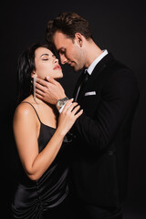 passionate woman standing with closed eyes near elegant man touching her neck isolated on black