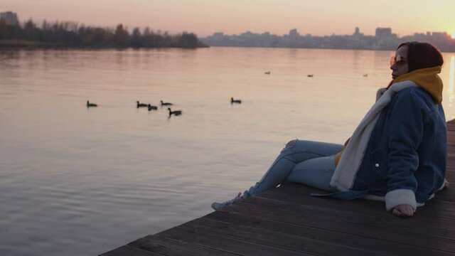 Side view of young woman sitting on pier in evening. Female rests and looks at water with ducks in urban environment.