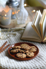 Plate of chocolate chip cookies, cinnamon sticks, soft blanket, open book and lit candles. Hygge at home. Selective focus.
