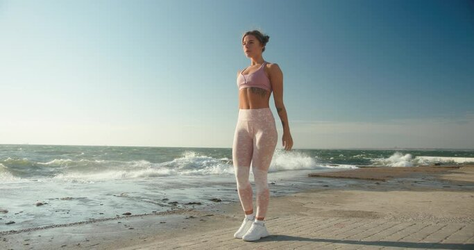 Young blonde woman with ponytail in pink top leggings and sneakers exercises on embankment against high storm sea waves slow motion