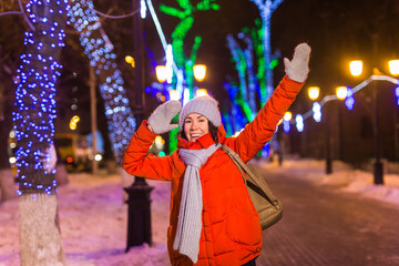 Girl having fun on christmas decoration lights street. Young happy smiling woman wearing stylish knitted scarf and jacket outdoors. Model laughing. Winter wonderland city scene, New Year party.
