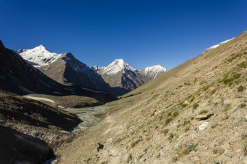 A view of snow capped Himalayan mountains under a blue sky from a trekking trail on the steep slope of a hill in Zanskar.