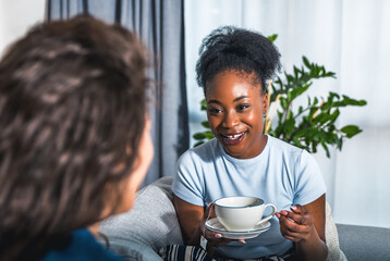 Two young women best friends sitting on the sofa at home drinking tea or coffee complaining on her...