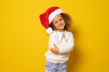 Beautiful smiling little girl in santa hat with arms folded over yellow background.