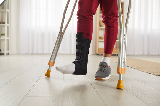 Unrecognizable person with broken leg or foot injury walking on crutches. Man wearing leg brace ankle support adjustable strap fracture fixator standing in living room. Cropped low section close up