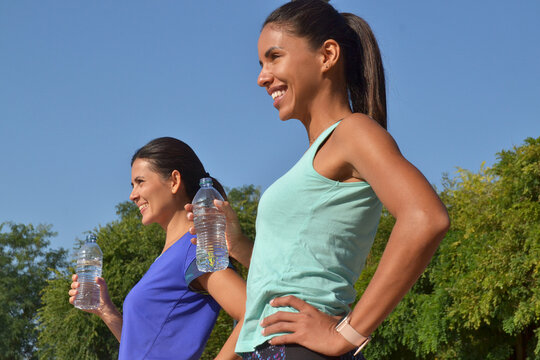 Two young latin women drinking water bottle after outdoors training on blue sky background.