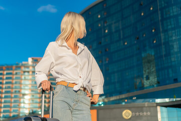 Portrait young blonde woman turned away from the camera in a white shirt and jeans with a suitcase in her hand against the background of a modern glass hotel in the rays of the setting sun,copy space