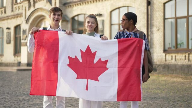 Happy Caucasian students posing with African American groupmate holding Canadian flag smiling looking at camera. Three smart confident women and men standing at university campus outdoors