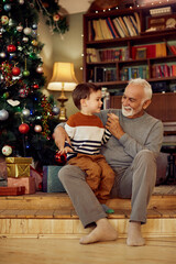 Obraz na płótnie Canvas Happy grandfather and grandson talk while sitting by Christmas tree at home.