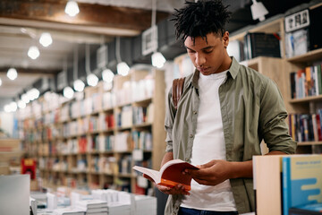 Young African American student reads book while standing at bookstore.