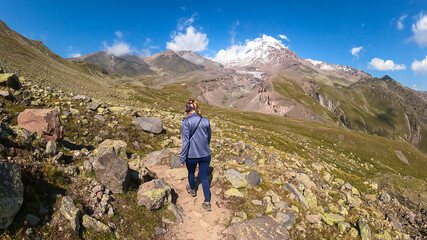 Naklejka na ściany i meble A woman walking on a narrow path towards Mount Kazbeg in Caucasus, Georgia. There slopes are overgrown with green grass. Massive Gergeti Glacier under the sharp peak. Cloudless sky. Exploring.