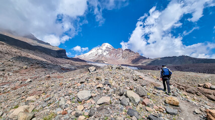 Naklejka premium A man crossing a stony slope towards Mount Kazbeg in Caucasus, Georgia. Barren slopes below the snow-capped peak and the Gergeti Glacier. Tranquillity. Natural remedy. Massive glacier foot. Few cloud
