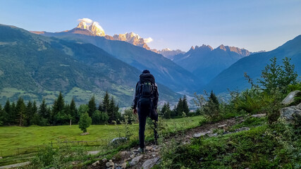 A man with big hiking backpack standing on a lush pasture with a view on the first sunbeams reaching the peaks of Ushba in Caucasus, Georgia. Cloudless sky above the high and snow-capped mountains.