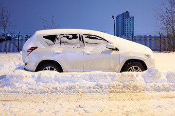 White car covered with snow at night. Parked car in the yard. City after snow storm. Side view.