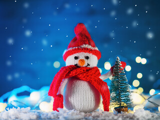 Funny knitted toy snowman. Christmas decor, fake snow decoration , bokeh garlands, blue background