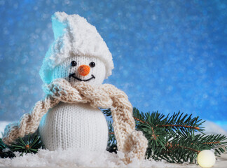 Cute knitted toy snowman in a hat and scarf in the snow, blue background. Christmas holiday...