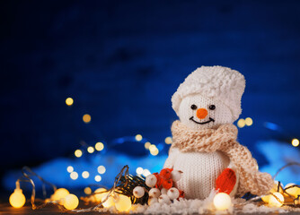 Obraz premium Funny knitted toy snowman. Christmas decor, fake snow decoration , bokeh garlands, blue background copy space