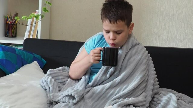 a boy with a cold is at home, being treated. there are medicines, medicine, throat and nose spray nearby. sick kid with runny nose and fever heat lying on couch at home. drinking a hot drink