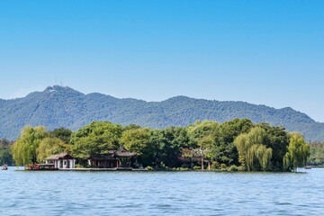 Fototapeta na wymiar A small green island on the West Lake in Hangzhou, China, filled with green trees and surrounded by hills, mountains and a blue sky