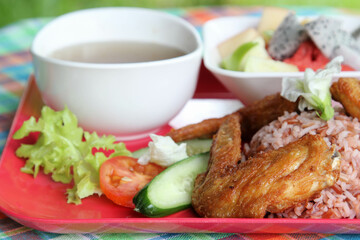 Thai style food, the deep fried chicken wings with rice, soup, vegetable and fruit on red plate.