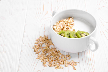 yogurt with kiwi and oatmeal in a white bowl and with oatmeal grains on a white wooden background